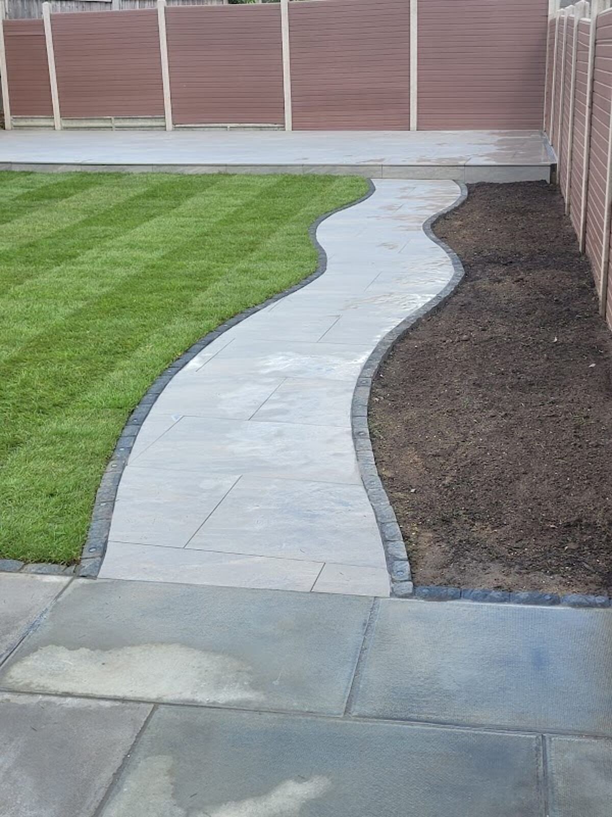 Curved porcelain garden path with dark edging between new turf and a planting bed